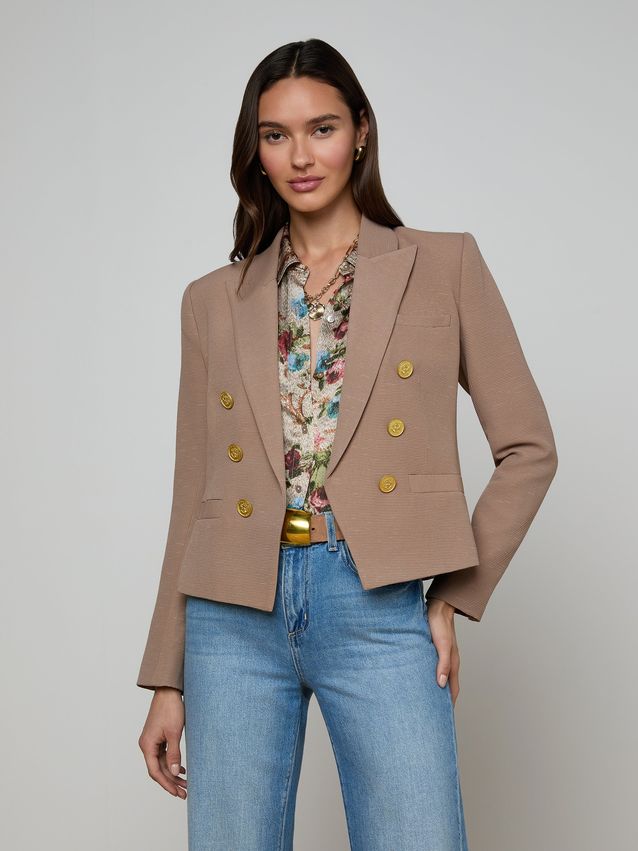 A woman stands against a plain background wearing the Brooke Blazer, an open-front double-breasted style with gold buttons, paired with a floral blouse and blue jeans. Her hand rests in her pocket and she is smiling slightly.