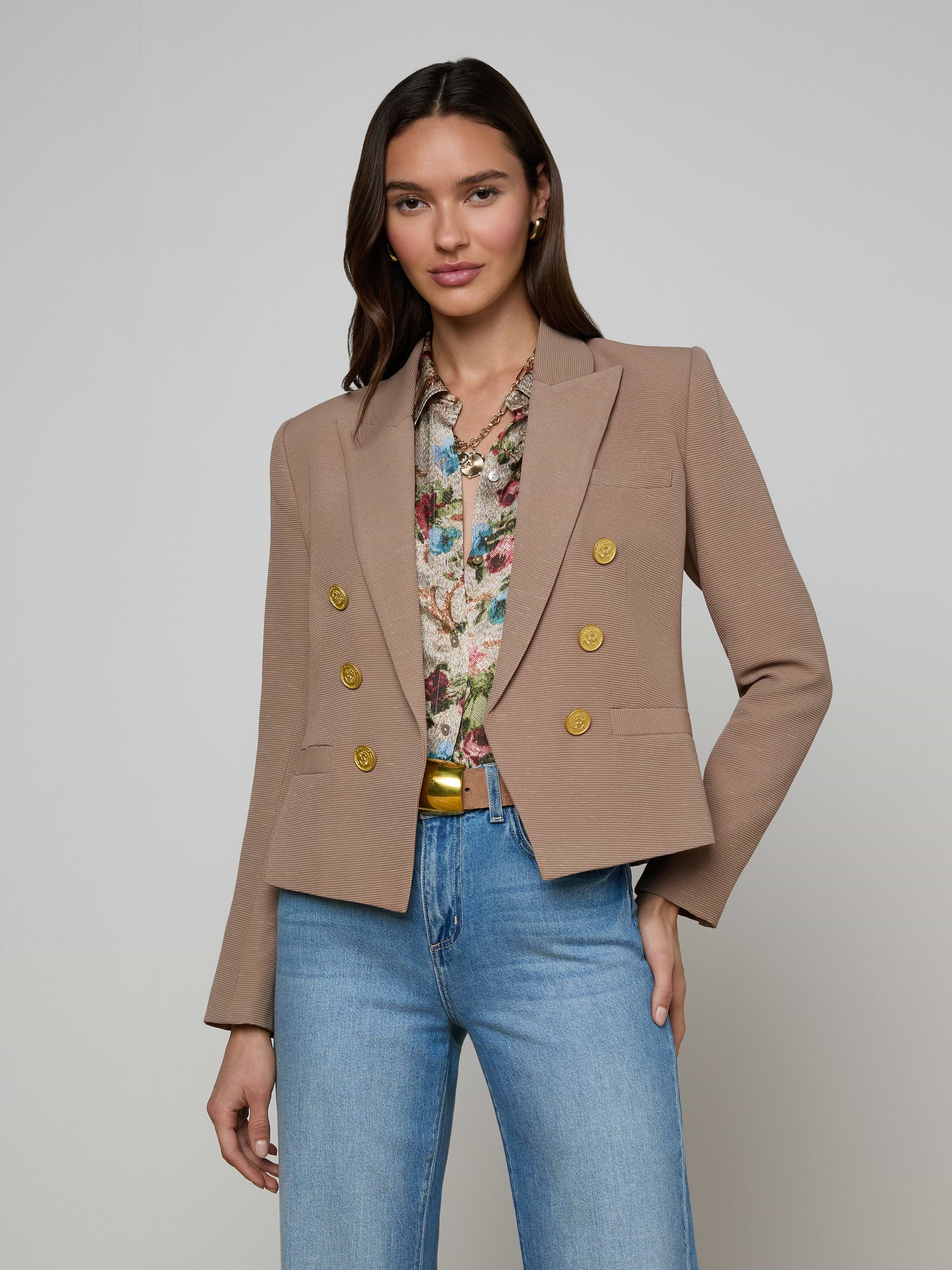 A woman stands against a plain background wearing the Brooke Blazer, an open-front double-breasted style with gold buttons, paired with a floral blouse and blue jeans. Her hand rests in her pocket and she is smiling slightly.