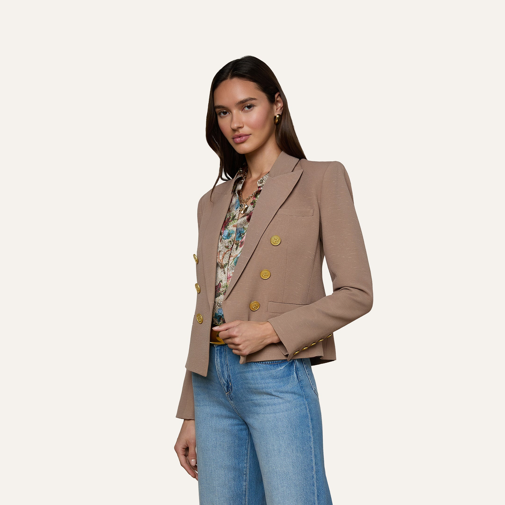 A woman wearing the Brooke Blazer—a beige, double-breasted blazer with gold buttons crafted from lightweight ribbed fabric—poses against a plain light background, styled with a floral blouse and blue jeans.