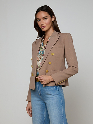 A woman wearing the Brooke Blazer in light brown with gold buttons, paired with a floral blouse and light blue jeans, stands against a plain background. The blazer’s lightweight ribbed fabric adds subtle texture to her classic style.