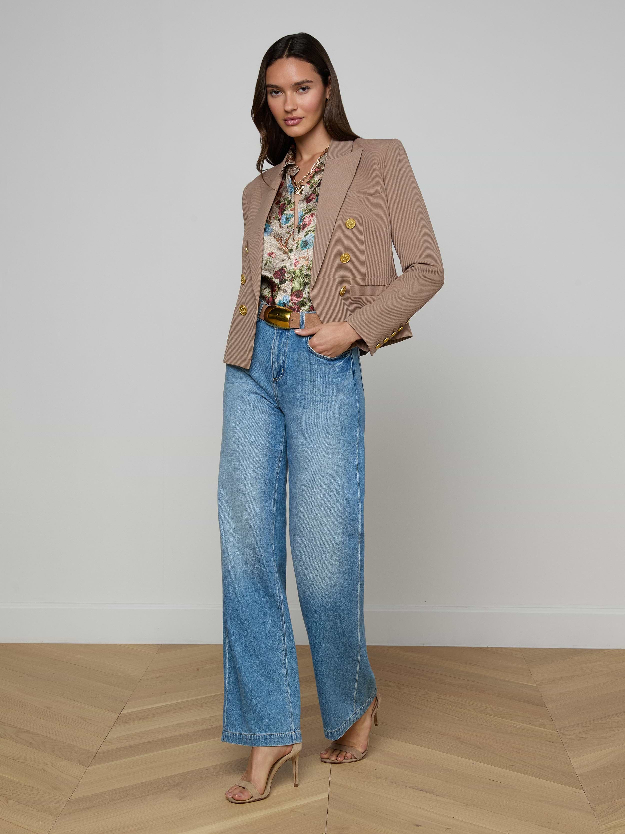 A woman stands against a plain background wearing the Brooke Blazer in tan, styled over a floral blouse with wide-leg blue jeans and beige heels, posing confidently with one hand in her pocket.