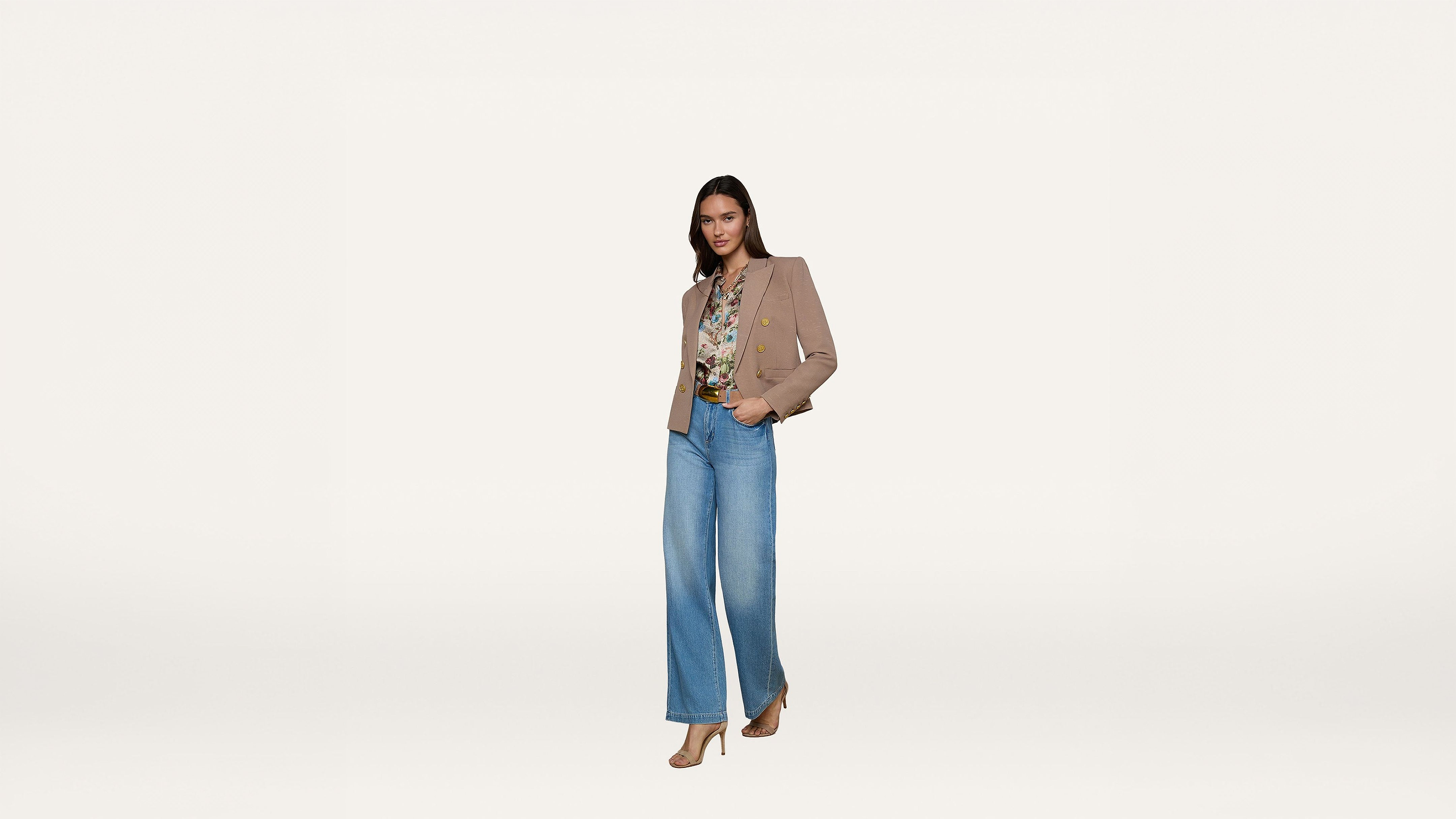 A woman stands confidently, wearing a tan blazer, printed blouse, wide-leg blue jeans, and beige heels against a plain white background.