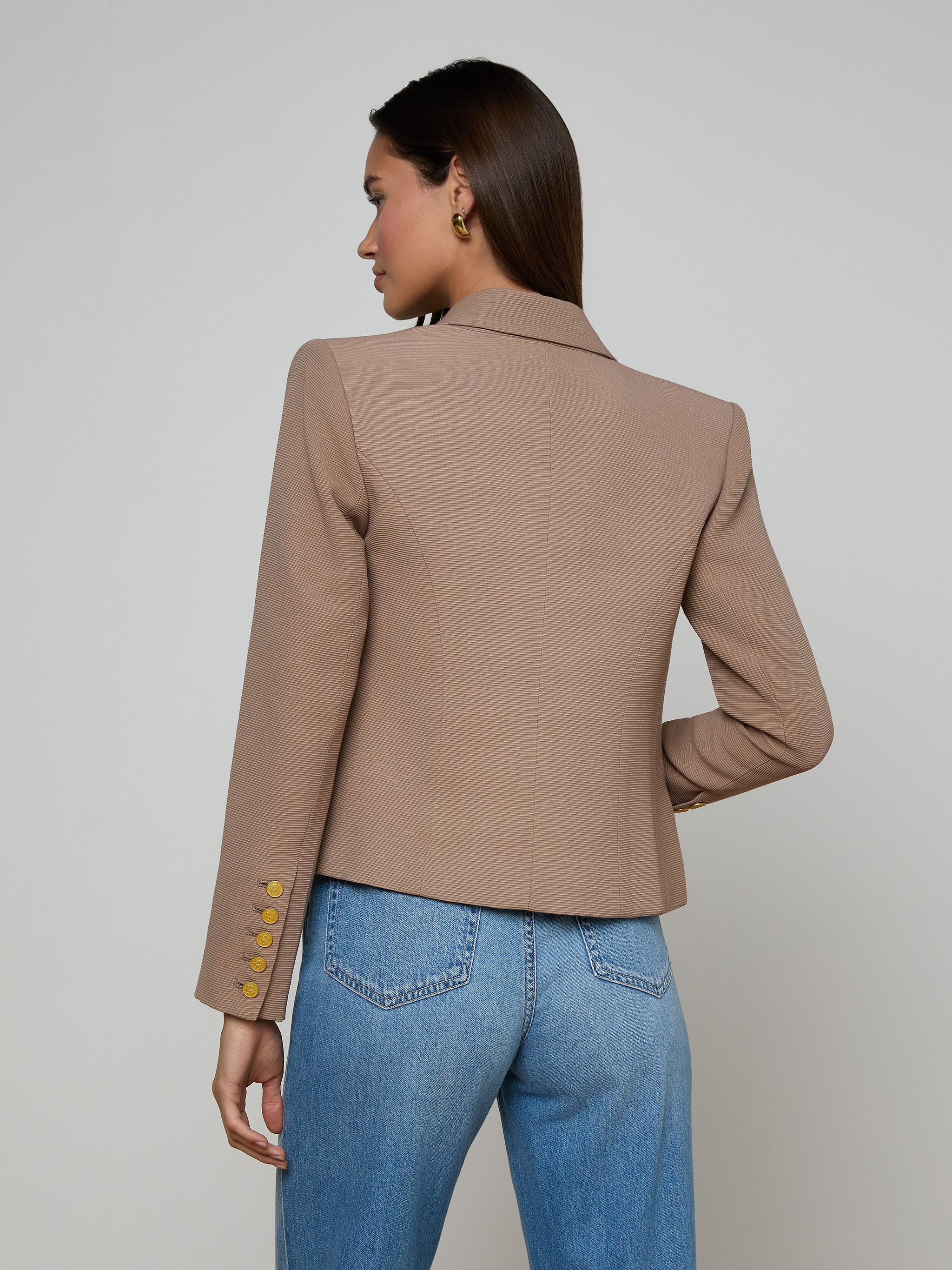 A woman stands facing away, wearing the Brooke Blazer—a tan double-breasted style featuring gold buttons on the sleeves—paired with blue jeans. Her dark hair is straight and tucked behind one ear, set against a plain, light-colored background.