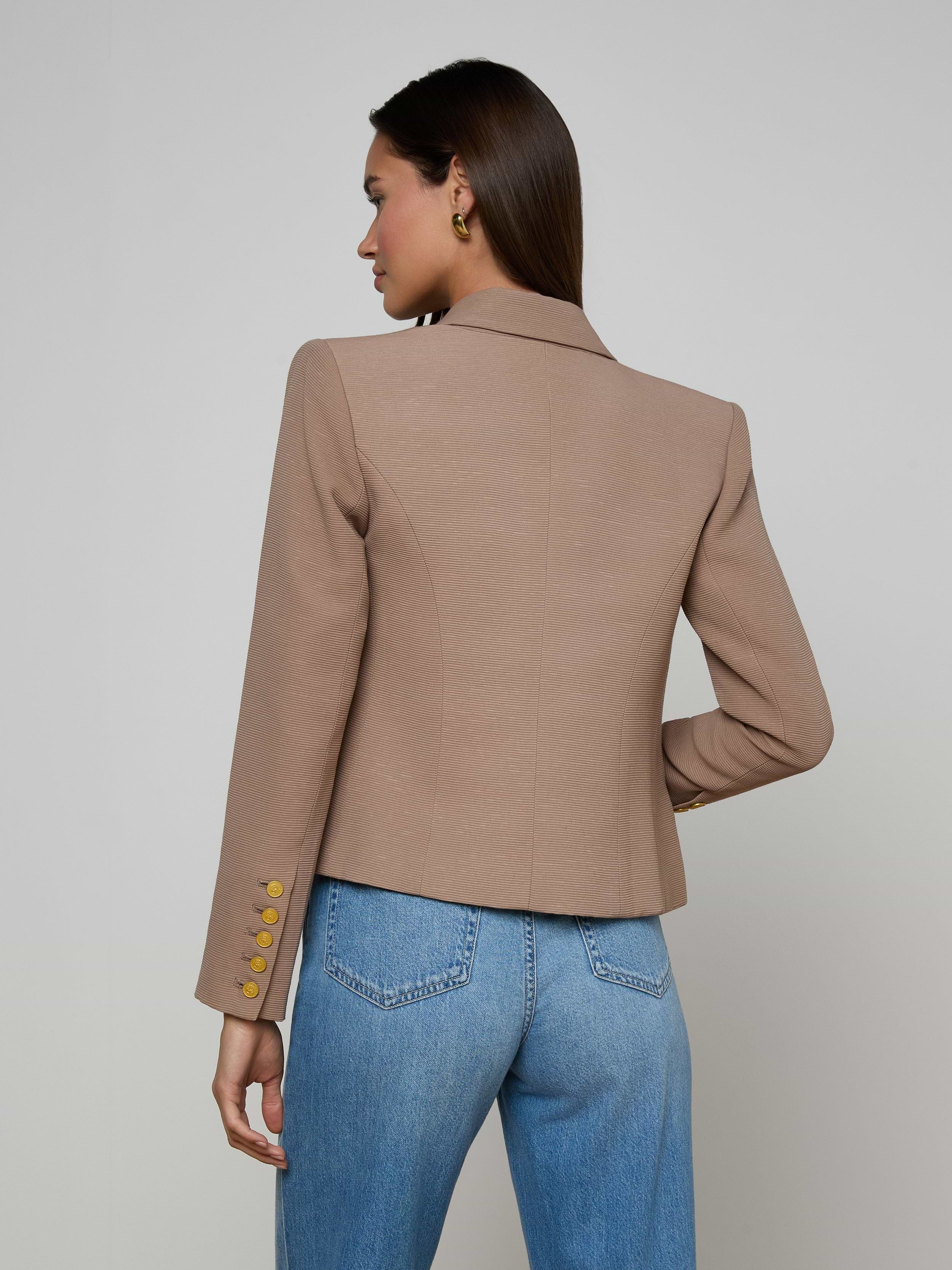 A woman stands facing away, wearing the Brooke Blazer—a tan double-breasted style featuring gold buttons on the sleeves—paired with blue jeans. Her dark hair is straight and tucked behind one ear, set against a plain, light-colored background.