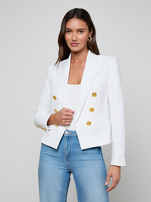 A woman with long brown hair wears the Brooke Blazer—a white open-front blazer with gold buttons—over a white top and light blue jeans, standing against a plain light gray background.