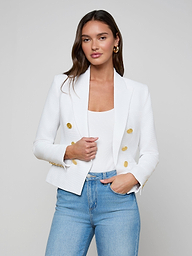 A woman stands against a plain light background with one hand in her pocket, wearing the Brooke Blazer—a white double-breasted blazer with gold buttons—paired with a white top and light blue jeans.