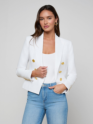 A woman with long brown hair wears the Brooke Blazer, an open-front white blazer with gold buttons, over a white top and light blue jeans. She stands against a plain light background, looking at the camera with a neutral expression.