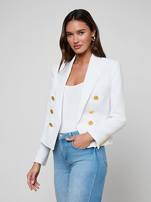A woman with long brown hair wears the Brooke Blazer—an open-front white blazer with gold buttons—over a white top and light blue jeans, standing against a plain light gray background.