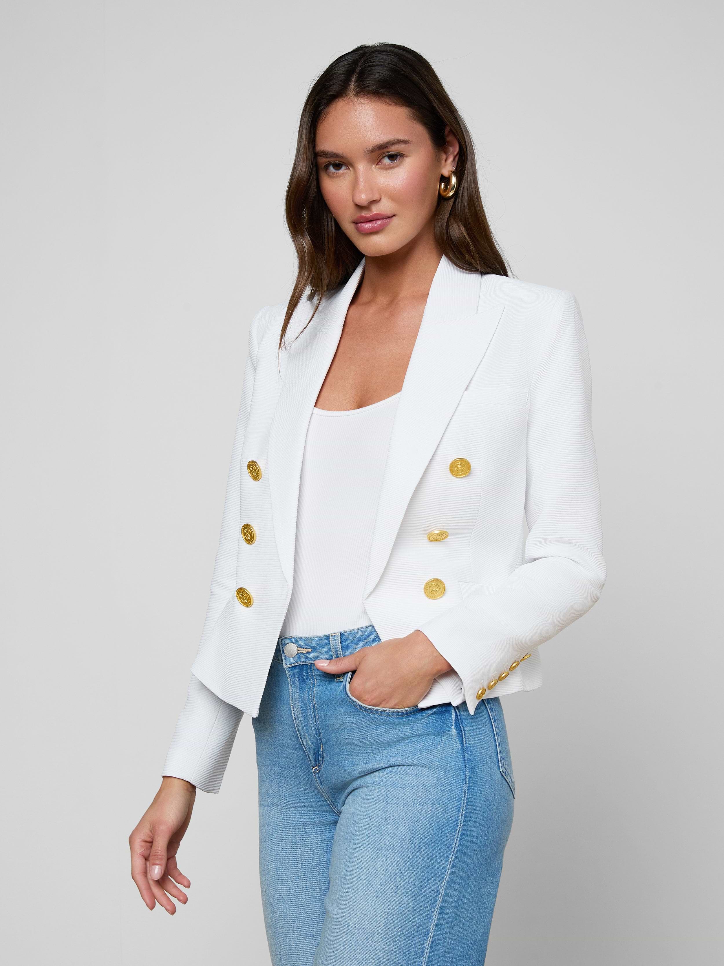 A woman wearing the Brooke Blazer—a double-breasted white blazer with gold buttons—paired with a white top and light blue jeans, stands against a plain light background, looking at the camera with a slight smile.