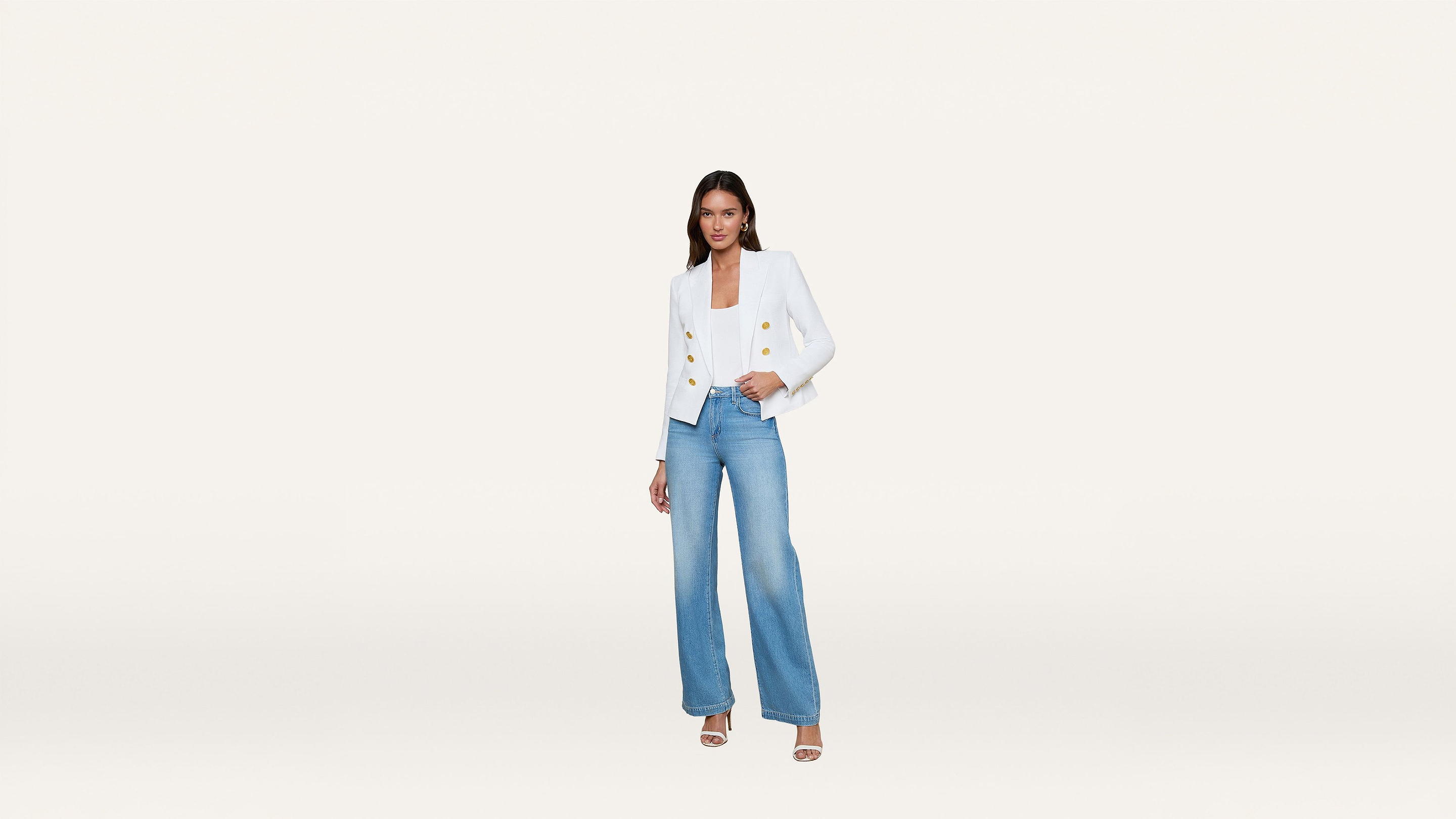 Woman wearing a white blazer with gold buttons, blue wide-leg jeans, and white heels, standing against a plain white background.