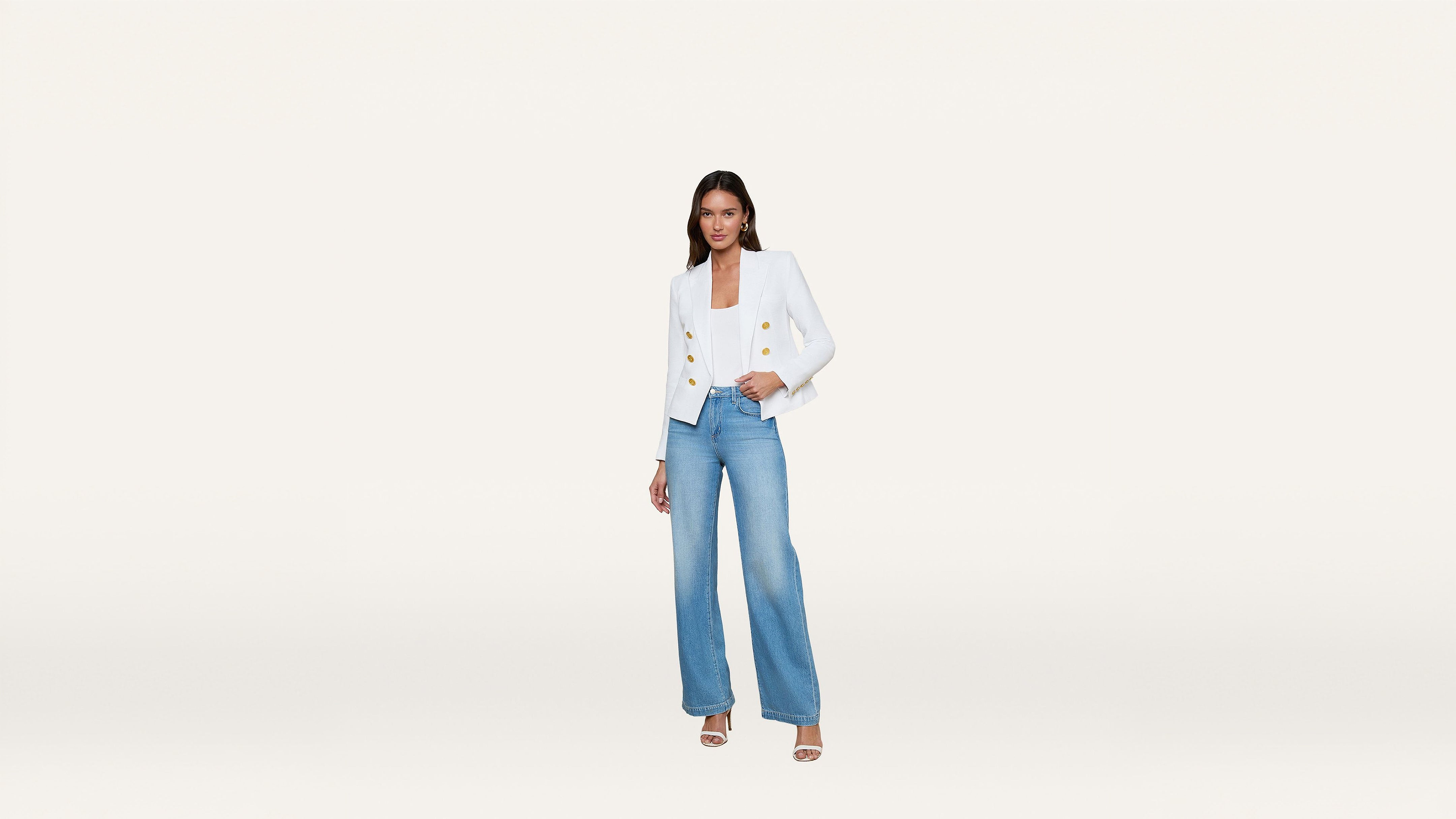 Woman wearing a white blazer with gold buttons, blue wide-leg jeans, and white heels, standing against a plain white background.