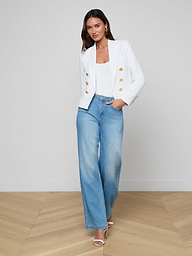 A woman stands against a plain background wearing the Brooke Blazer—a double-breasted white blazer with gold buttons—paired with a white top, wide-leg blue jeans, hoop earrings, and heeled sandals.