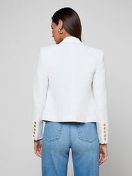 A woman with straight brown hair and gold hoop earrings stands facing away, wearing the Brooke Blazer—a lightweight ribbed white blazer with gold buttons on the sleeves—paired with light blue high-waisted jeans, set against a plain light background.