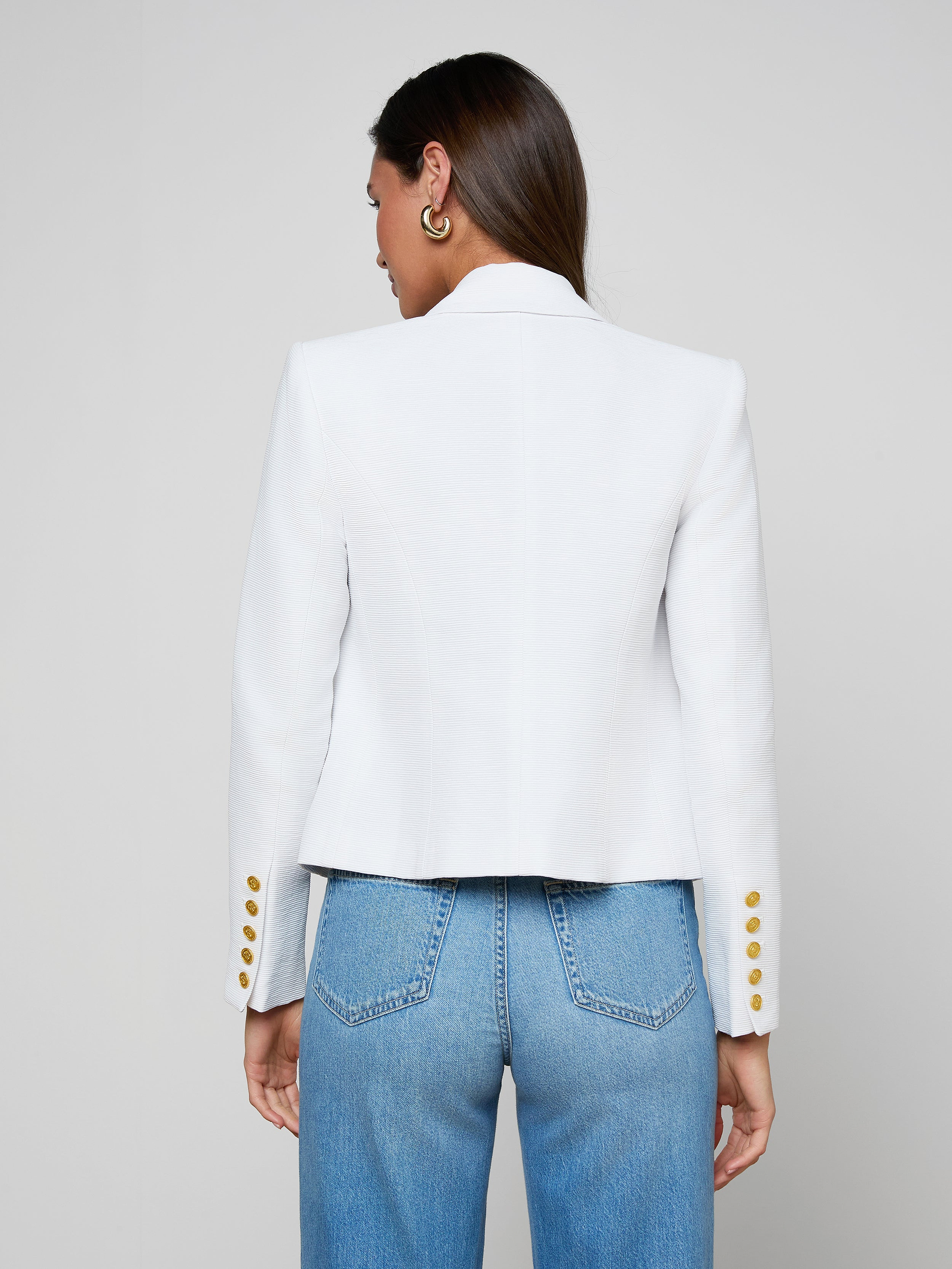 A woman with straight brown hair and gold hoop earrings stands facing away, wearing the Brooke Blazer—a lightweight ribbed white blazer with gold buttons on the sleeves—paired with light blue high-waisted jeans, set against a plain light background.