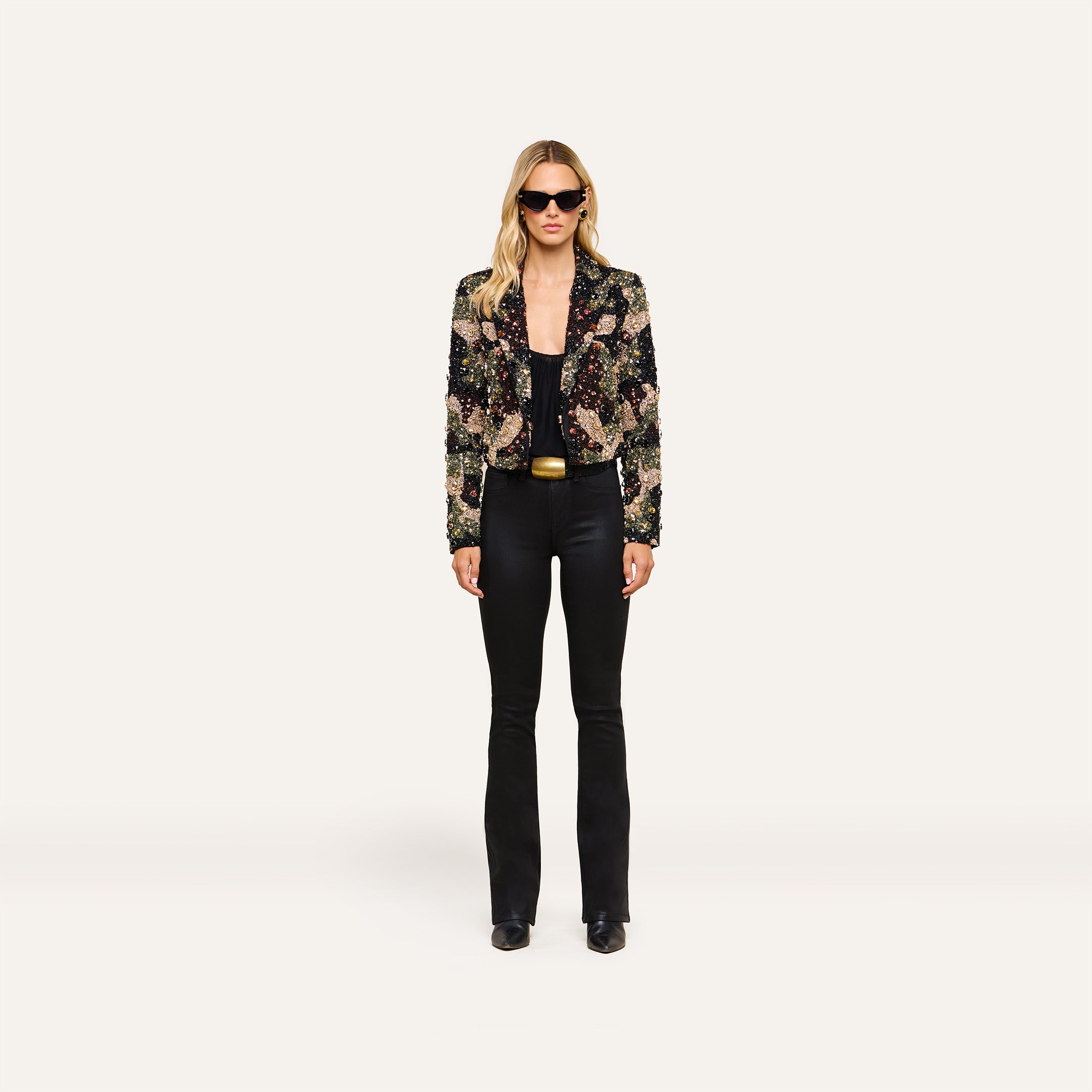 Woman wearing black sunglasses, the Jen Crystal Blazer—a couture-inspired, hand-embellished piece—paired with a black top, black flared pants, and black shoes stands against a plain white background.