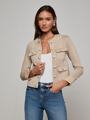 A woman with long brown hair models the L'AGENCE Yari Jean Jacket over a white top, standing against a light gray background. She holds one side of the jacket with her left hand and smiles slightly.