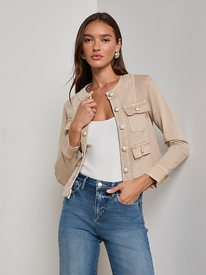 A woman with long brown hair wears the L'AGENCE Yari Jean Jacket, styled over a white top and blue jeans, standing confidently against a plain light wall.