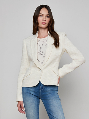 A woman with long brown hair wears the Jennah Tweed Blazer over a patterned top and blue jeans, standing against a light gray background with one hand on her hip and a neutral expression.