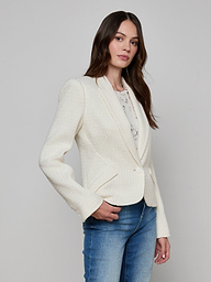 A woman with long brown hair is wearing the Jennah Tweed Blazer in ivory over a light shirt and blue jeans, standing against a plain off-white background and looking slightly to the side.