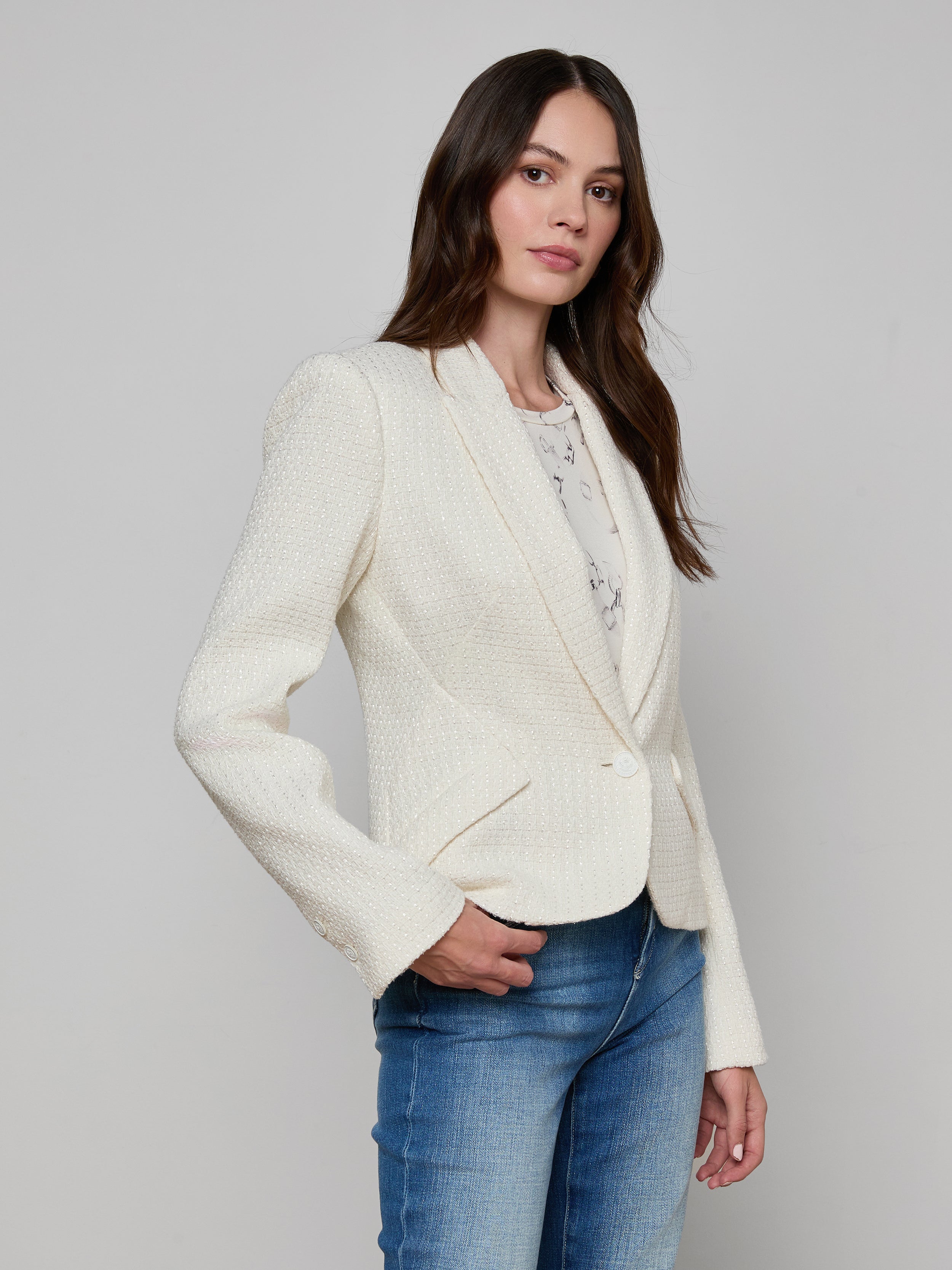 A woman with long brown hair is wearing the Jennah Tweed Blazer in ivory over a light shirt and blue jeans, standing against a plain off-white background and looking slightly to the side.