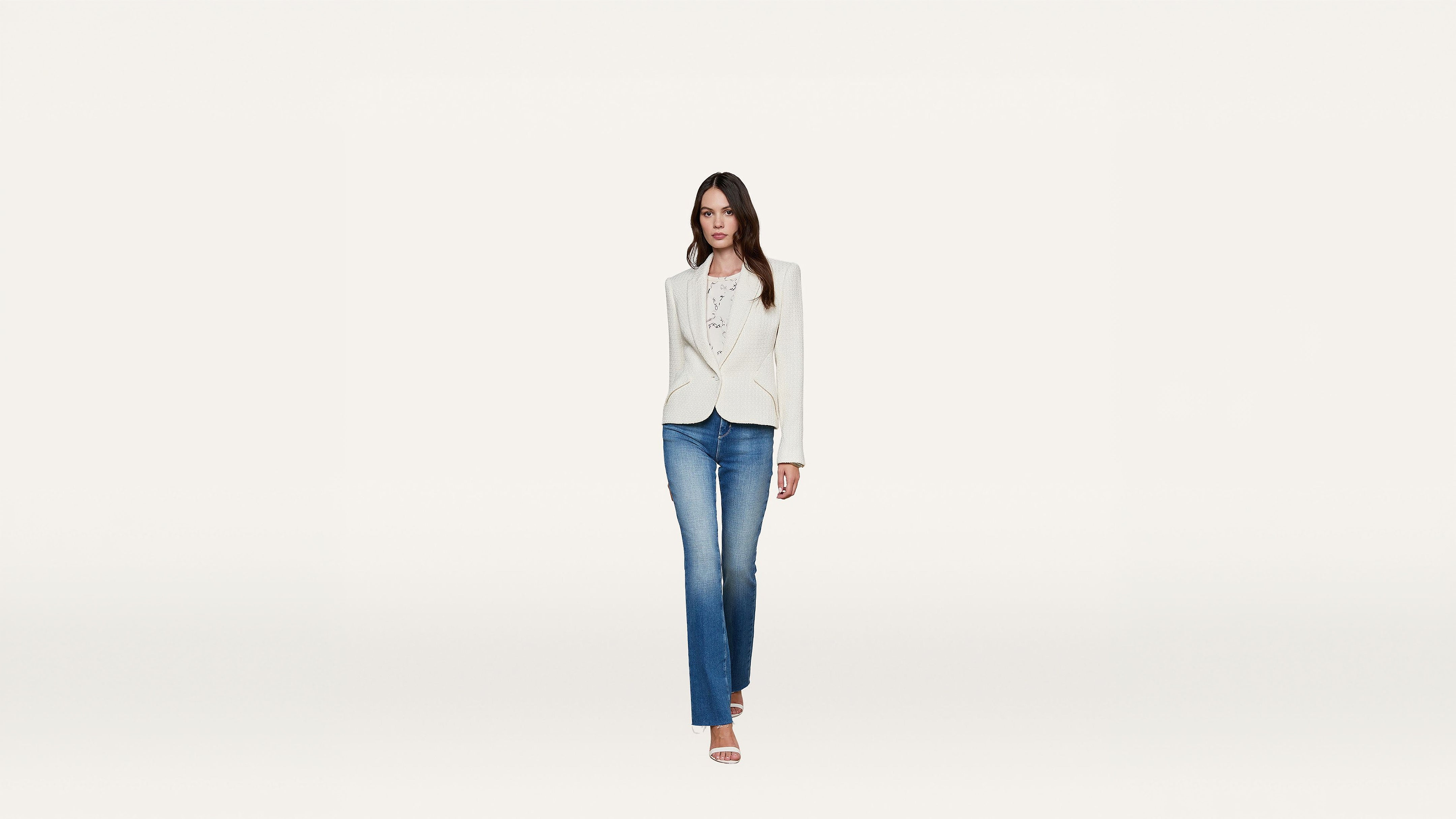 Woman walking forward wearing a white blazer, white top, blue jeans, and heeled sandals against a plain white background.