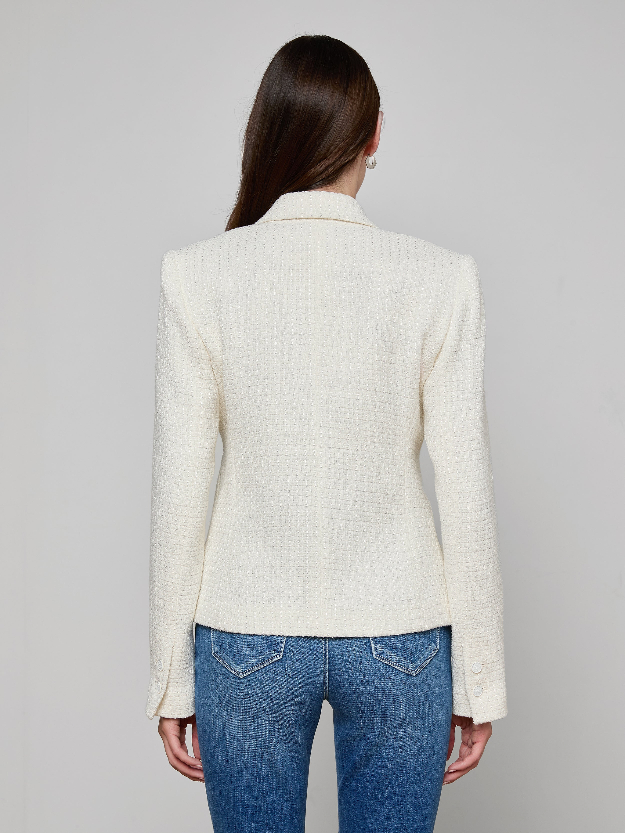 A woman with long brown hair faces away, wearing the petite Jennah Tweed Blazer in ivory and blue jeans against a plain light background.