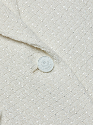 Close-up of the textured cream-colored fabric of the Jennah Tweed Blazer, showcasing a subtle grid pattern and a distinctive white button embellished with text and a decorative emblem.