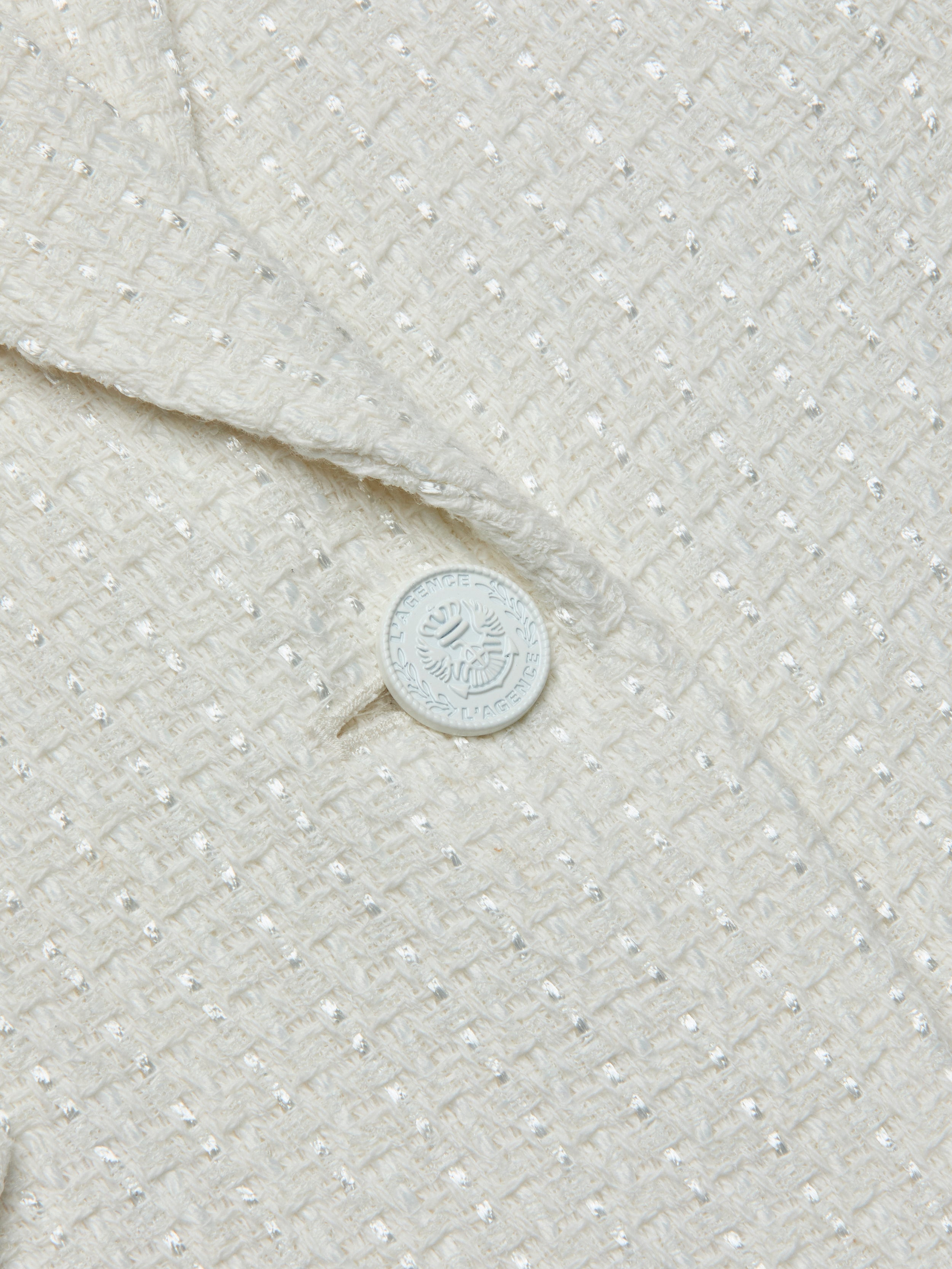 Close-up of the textured cream-colored fabric of the Jennah Tweed Blazer, showcasing a subtle grid pattern and a distinctive white button embellished with text and a decorative emblem.