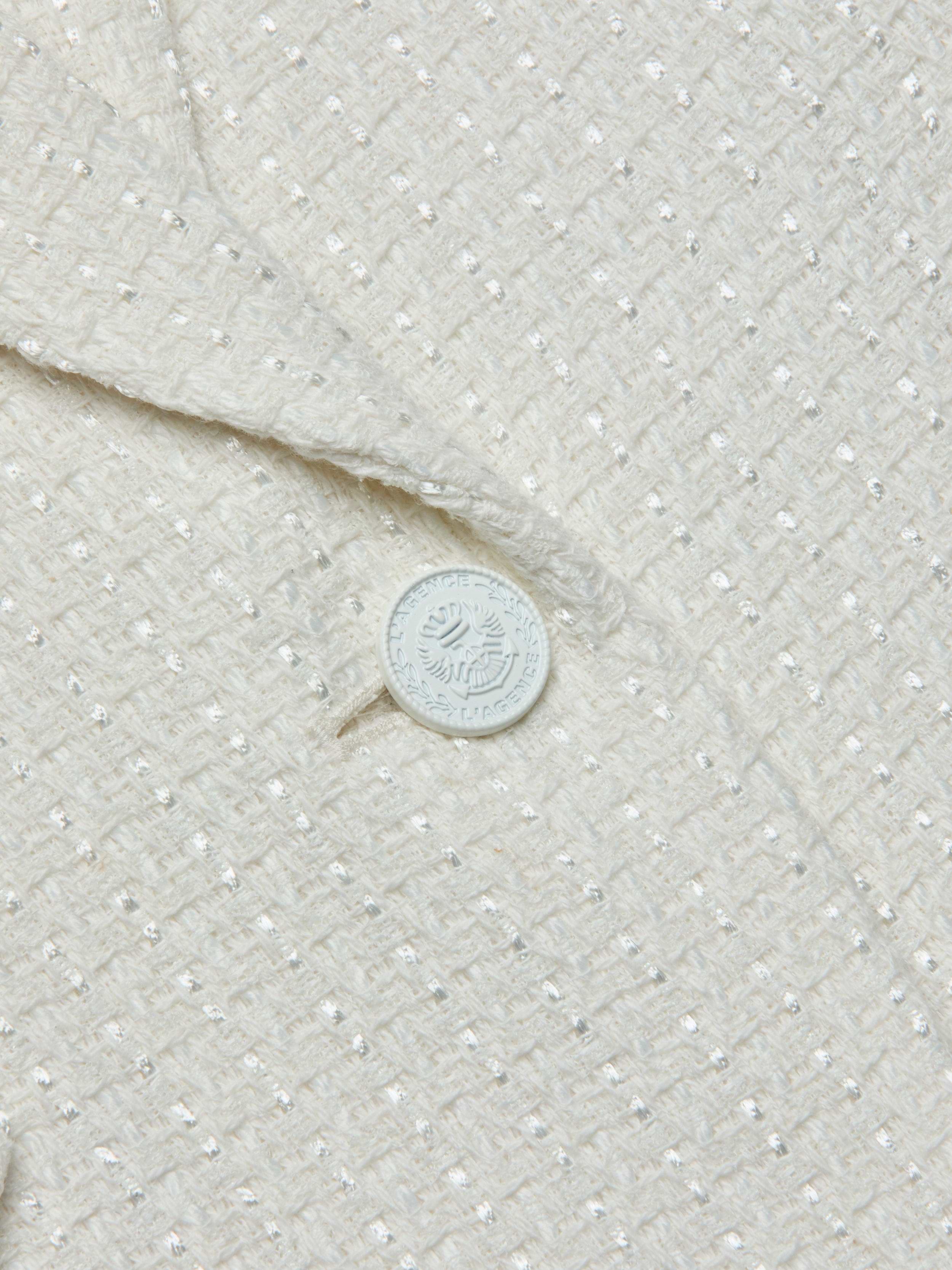 Close-up of the textured cream-colored fabric of the Jennah Tweed Blazer, showcasing a subtle grid pattern and a distinctive white button embellished with text and a decorative emblem.