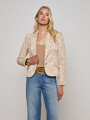 A woman with long blonde hair wears the L'AGENCE Sale Jennah Jacquard Blazer over a tan top and blue jeans with a wide brown belt and gold bracelet, standing against a plain light background.