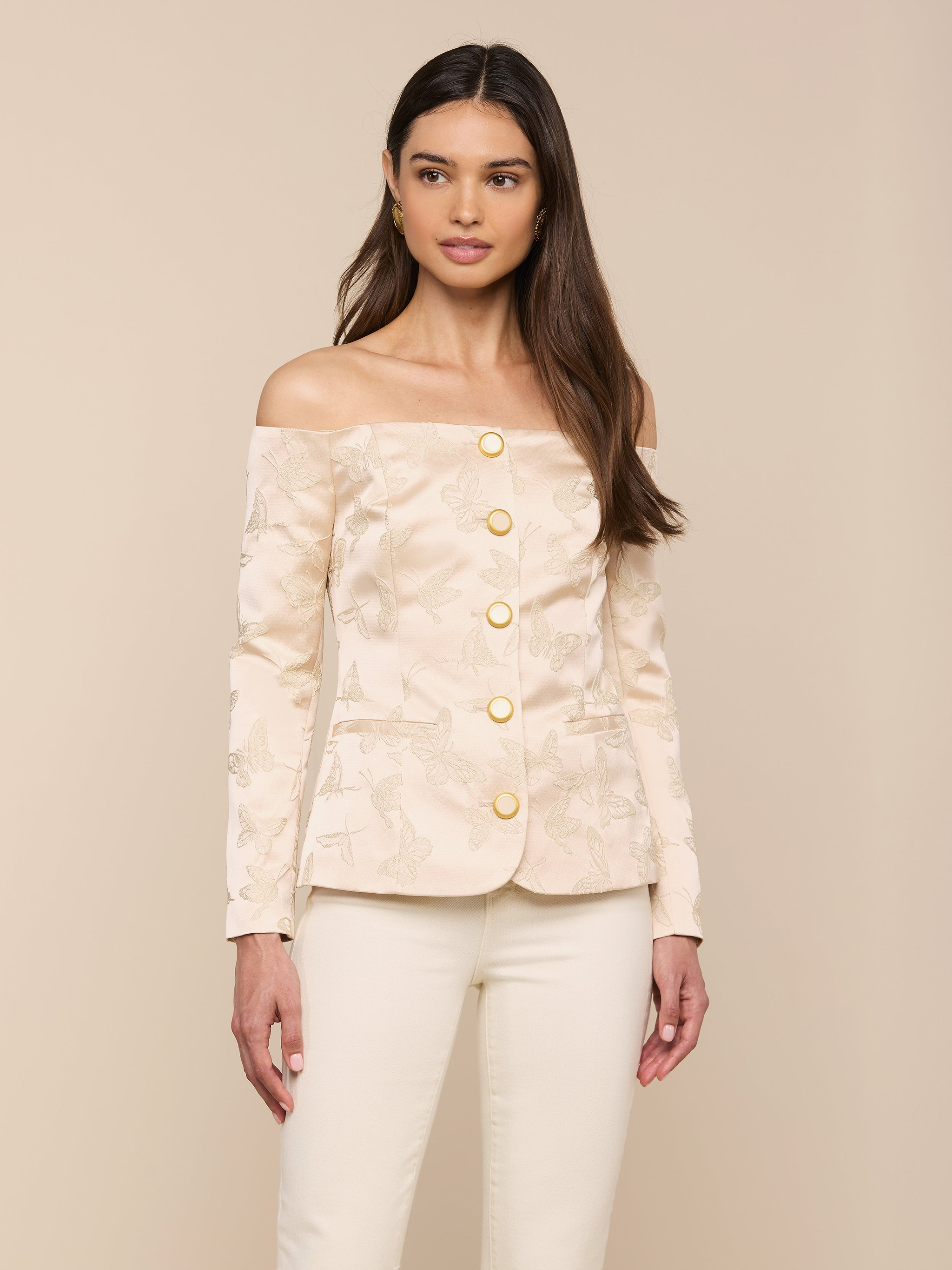 A woman stands against a plain light background, wearing the Alice Blazer Top—an off-the-shoulder cream blazer with gold buttons—paired with matching cream tailored suiting pants.