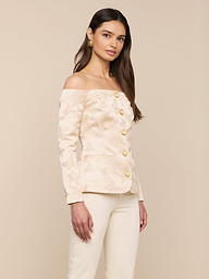 A woman stands against a plain background, wearing the Alice Blazer Top—a cream-colored, off-the-shoulder tailored suiting piece with large gold buttons—paired with matching cream pants. She has long brown hair and is accessorized with gold earrings.