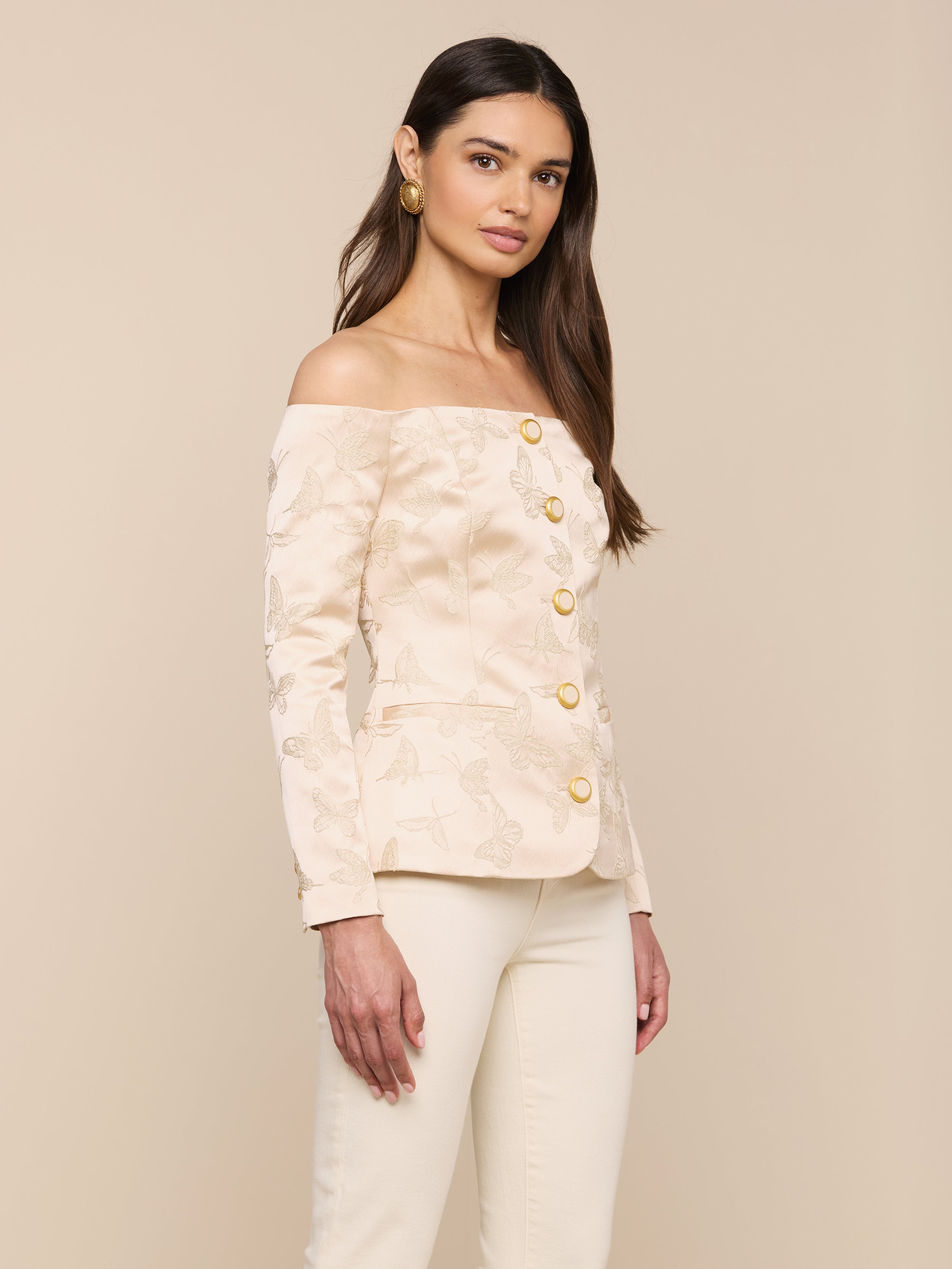 A woman stands against a plain background, wearing the Alice Blazer Top—a cream-colored, off-the-shoulder tailored suiting piece with large gold buttons—paired with matching cream pants. She has long brown hair and is accessorized with gold earrings.