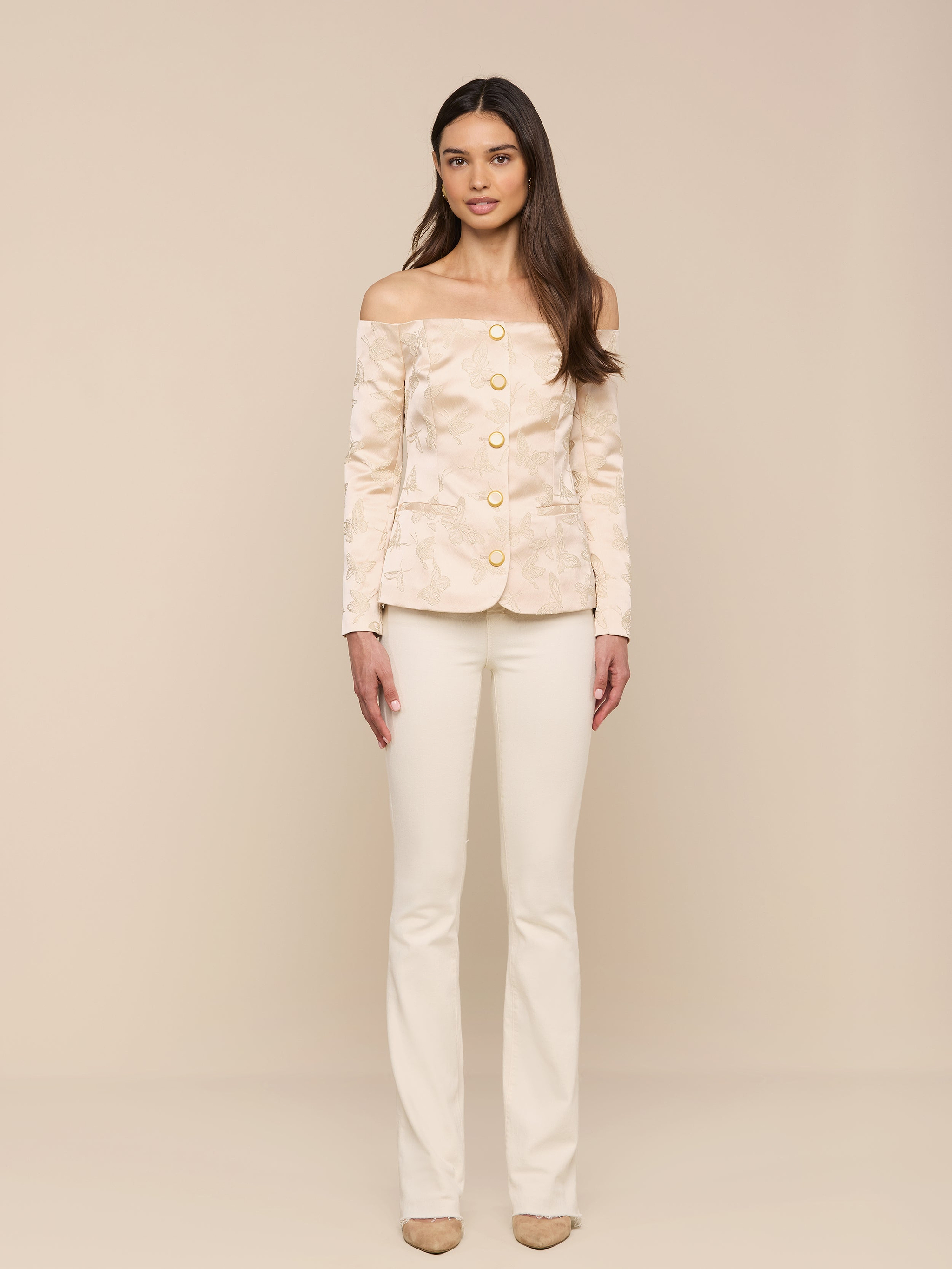 A woman stands facing forward, wearing the Alice Blazer Top—an off-the-shoulder beige suit with large buttons—paired with matching beige pants and shoes, against a plain white background.