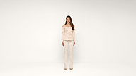 A woman stands facing forward in a minimal, white studio, wearing a light-colored, off-the-shoulder blouse and matching pants with heels.
