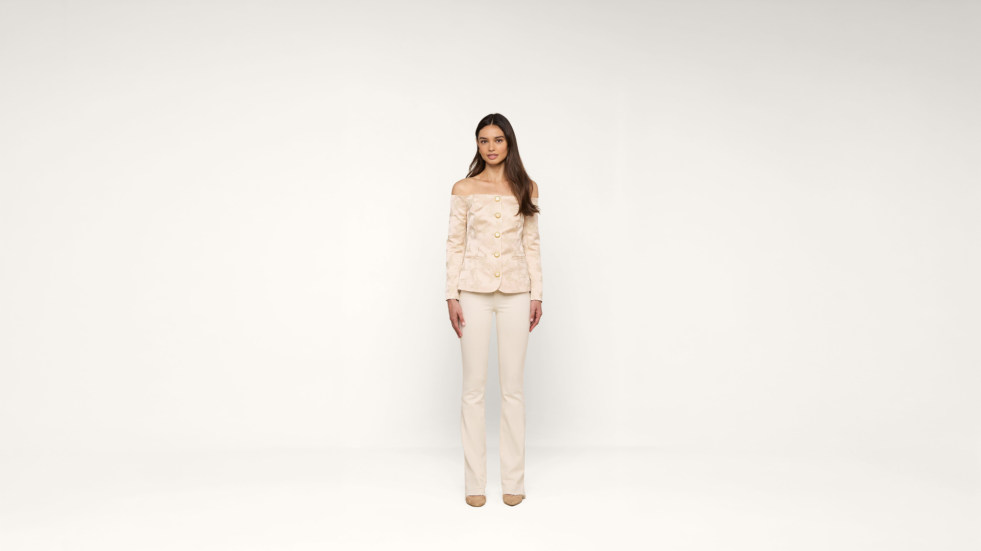 A woman stands facing forward in a minimal, white studio, wearing a light-colored, off-the-shoulder blouse and matching pants with heels.