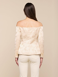 Une femme aux longs cheveux bruns, vue de dos, porte le Alice Blazer Top - un chemisier crème à motifs floraux, décolleté, avec des boutons de manche dorés - et un pantalon crème sur un fond neutre.