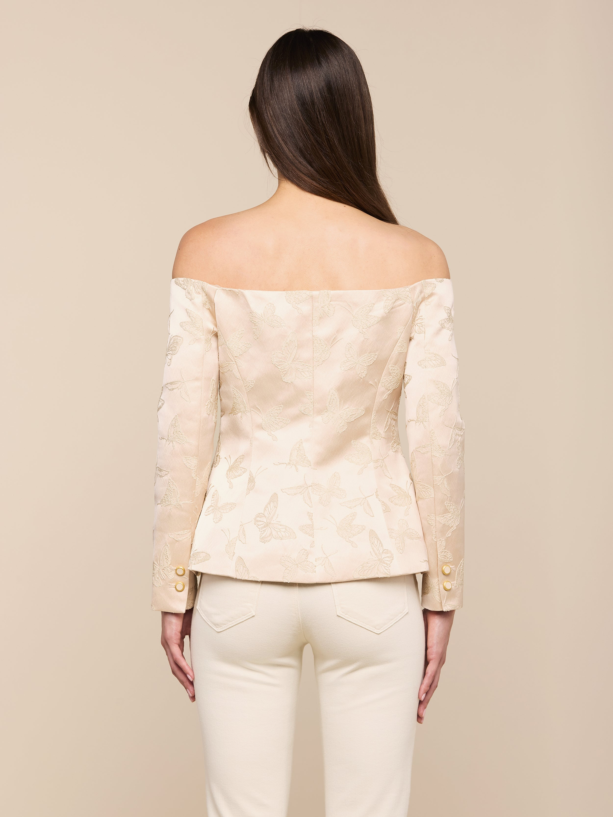 Une femme aux longs cheveux bruns, vue de dos, porte le Alice Blazer Top - un chemisier crème à motifs floraux, décolleté, avec des boutons de manche dorés - et un pantalon crème sur un fond neutre.