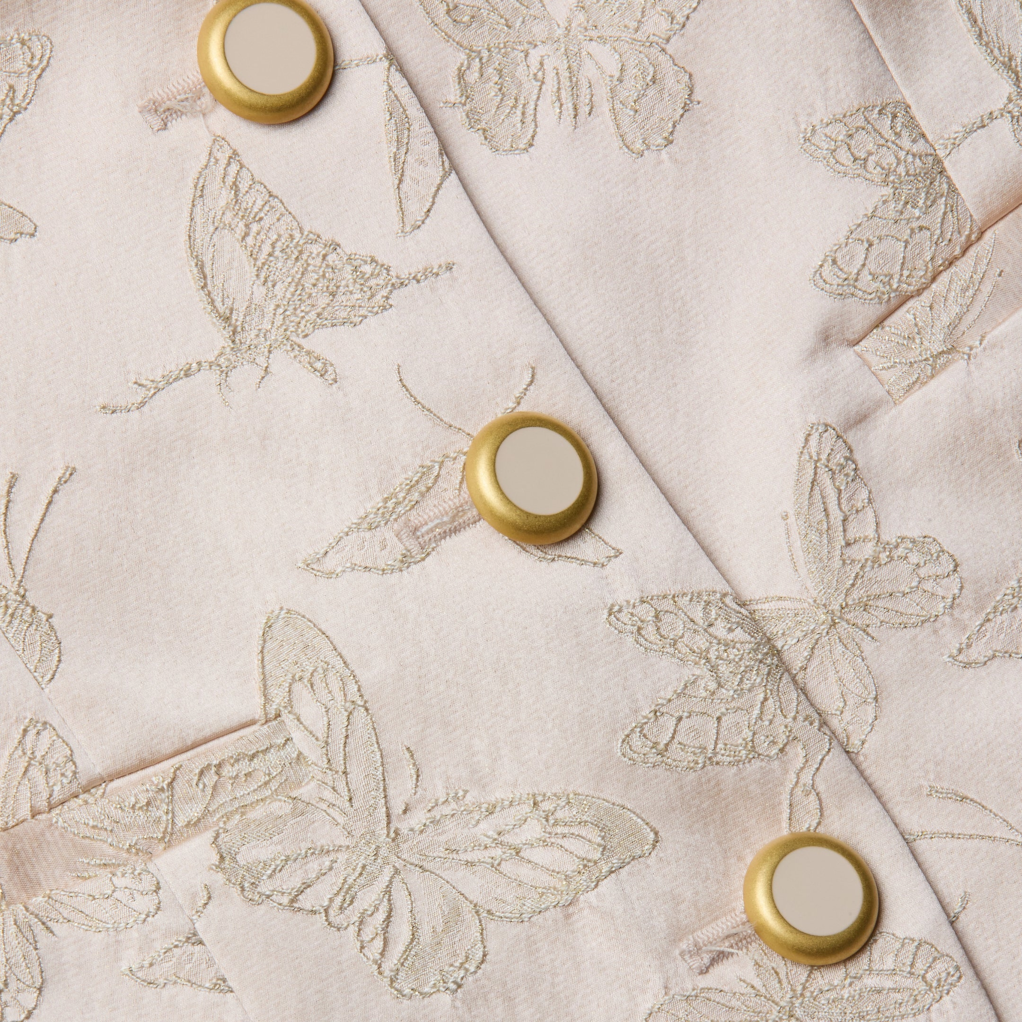Un gros plan du haut blazer Alice montre un tissu jacquard rose pâle avec des papillons brodés et des boutons dorés au centre crème, mettant en valeur les coutures détaillées et la texture lisse du satin.
