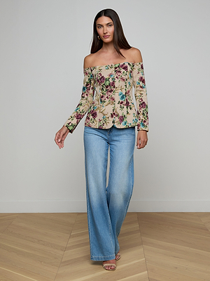 A woman stands on a wooden floor against a light wall, wearing the Alice Blazer Top with an off-the-shoulder floral design, paired with light blue wide-leg jeans and strappy sandals.