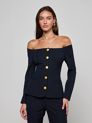 A woman exudes confidence in the L'AGENCE Alice Blazer Top, featuring an off-the-shoulder pinstripe design with bold gold buttons, posed against a simple background and staring directly at the camera with a calm expression.
