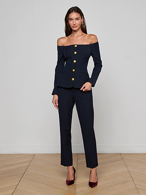 A woman poses indoors against a light wall in the L'AGENCE Alice Blazer Top—a navy, off-the-shoulder pinstripe suit with gold buttons—paired with matching pants, gold earrings, and burgundy heels for a chic power look.