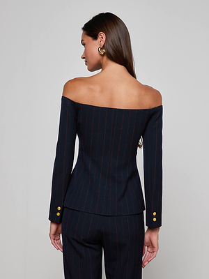 A woman faces away, exuding power in the L'AGENCE Alice Blazer Top—an off-the-shoulder pinstripe suit with gold sleeve buttons. Her long, loose hair is styled with large gold hoop earrings.
