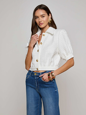 A woman with long brown hair poses against a plain background wearing the L'AGENCE Sale Cove Cropped Tweed Jacket with blue jeans, accessorized with large gold earrings and colorful bracelets.
