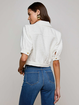 A woman with long brown hair and gold hoop earrings, wearing the L'AGENCE Sale Cove Cropped Tweed Jacket and blue high-waisted jeans, stands facing away from the camera.