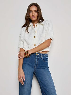 A woman with long brown hair and multiple bracelets wears the L'AGENCE Sale Cove Cropped Tweed Jacket, a white short-sleeved button-up, paired with high-waisted blue jeans against a plain background.