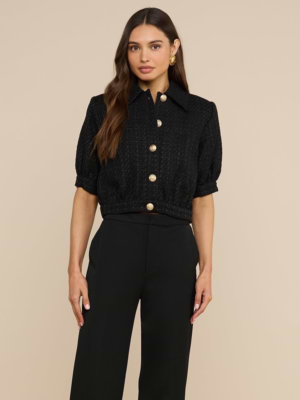 A woman stands against a plain light background wearing the Cove Cropped Tweed Jacket, a black short-sleeve button-up piece with gold buttons, paired with black pants and featuring a stylish cropped silhouette.