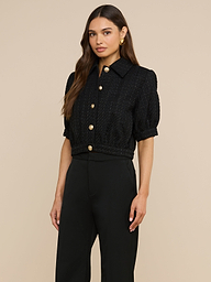 A woman stands against a plain background, wearing the Cove Cropped Tweed Jacket—a black collared short-sleeve jacket with large gold buttons and a cropped silhouette—paired with high-waisted black pants. She has long brown hair and accessorizes with gold earrings.