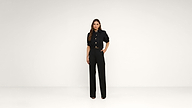 Woman stands against a plain white background, wearing a black button-up shirt and high-waisted black trousers with open-toe heels. One hand rests on her hip, and she looks straight ahead with a neutral expression.