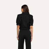 A woman with long brown hair stands facing away from the camera against a plain light background, wearing black pants and the Cove Cropped Tweed Jacket featuring a cropped silhouette.